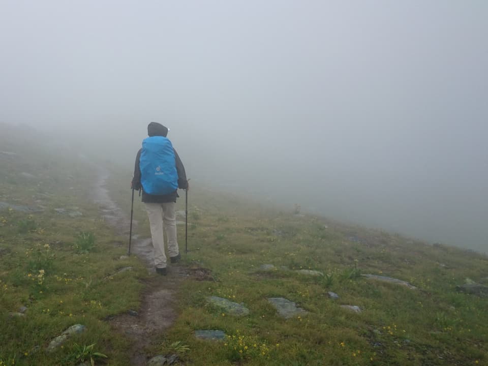 Diffuses Bild in den Nebel, knapp ist ein Wanderer von hinten erkennbar, auf grüner Wiese mit blauem Rucksack. 