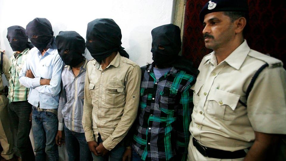Fünf verhaftete Männer mit schwarzen Tüchern mit Augenlöchern um den Kopf und ein Polizist.