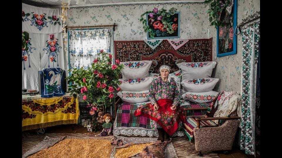 Eine ältere Frau sitzt in einem übbig mit Blumen und blumenmustern dekorierten Raum. Sie trägt ein buntes, traditionelles Kleid.