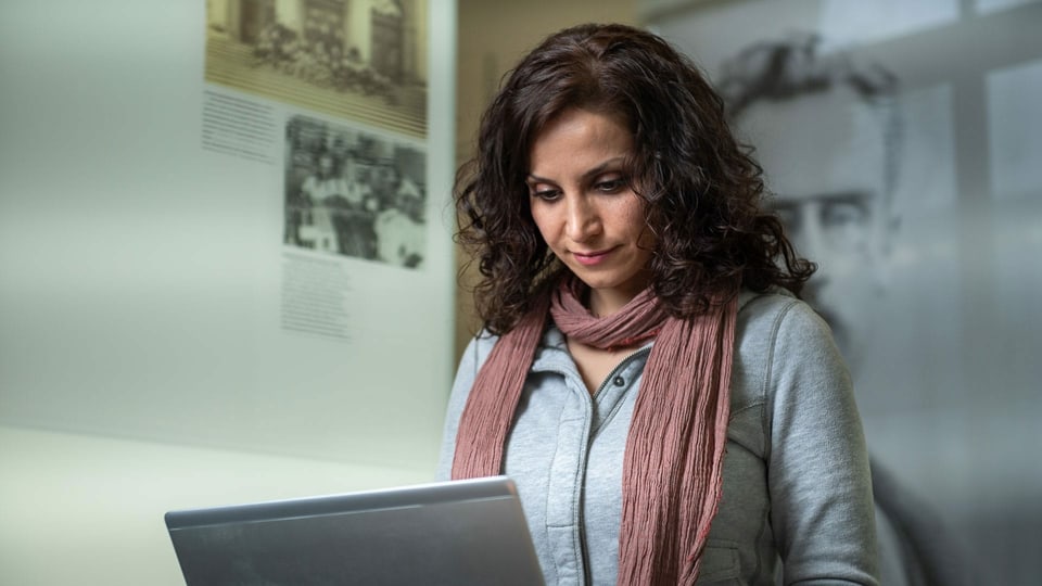 Eine junge Frau blickt vor einer Plakatwand in ihr Laptop.