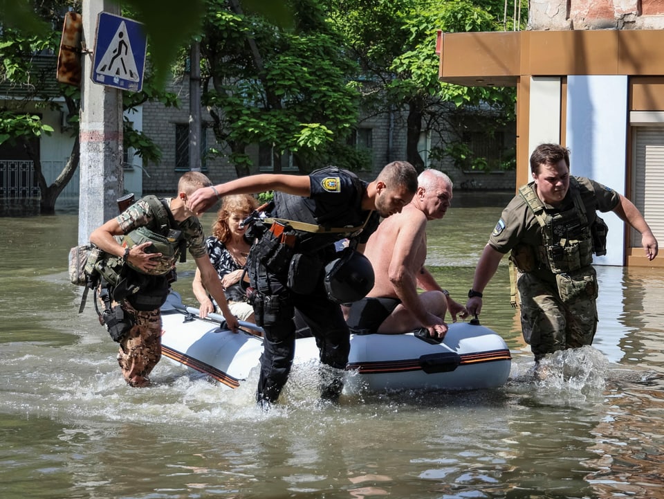 Drei Männer in Uniformen ziehen ein Schlauchboot durch Wasser in einer Stadt. Menschen sitzen im Boot.