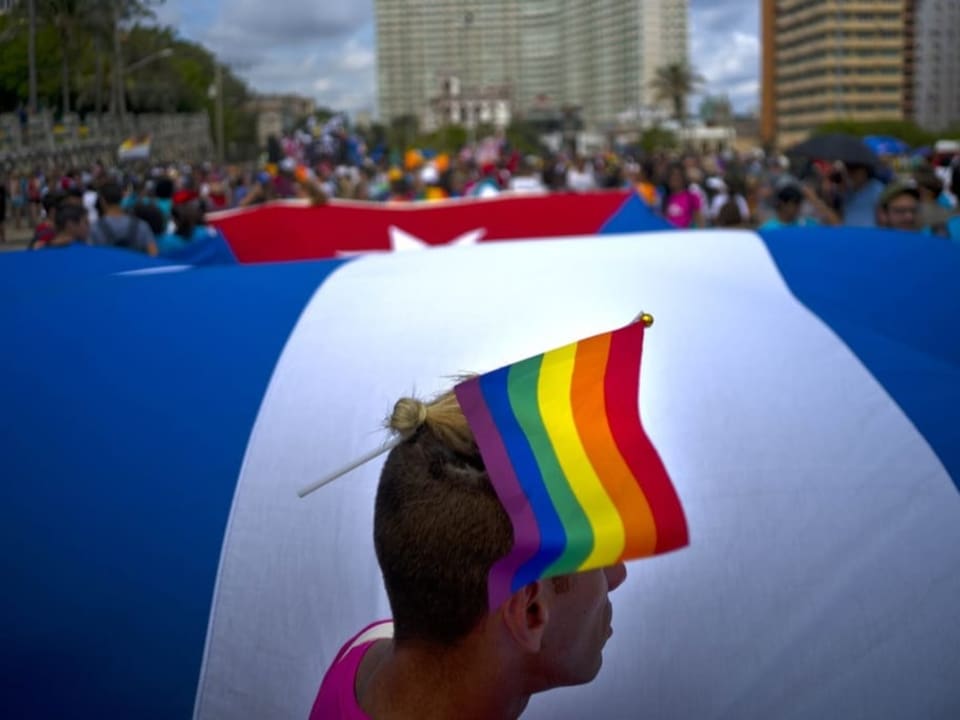 Mann vor kubanischer Riesenflagge, hat Regenbogenflagge im n den Haaren.