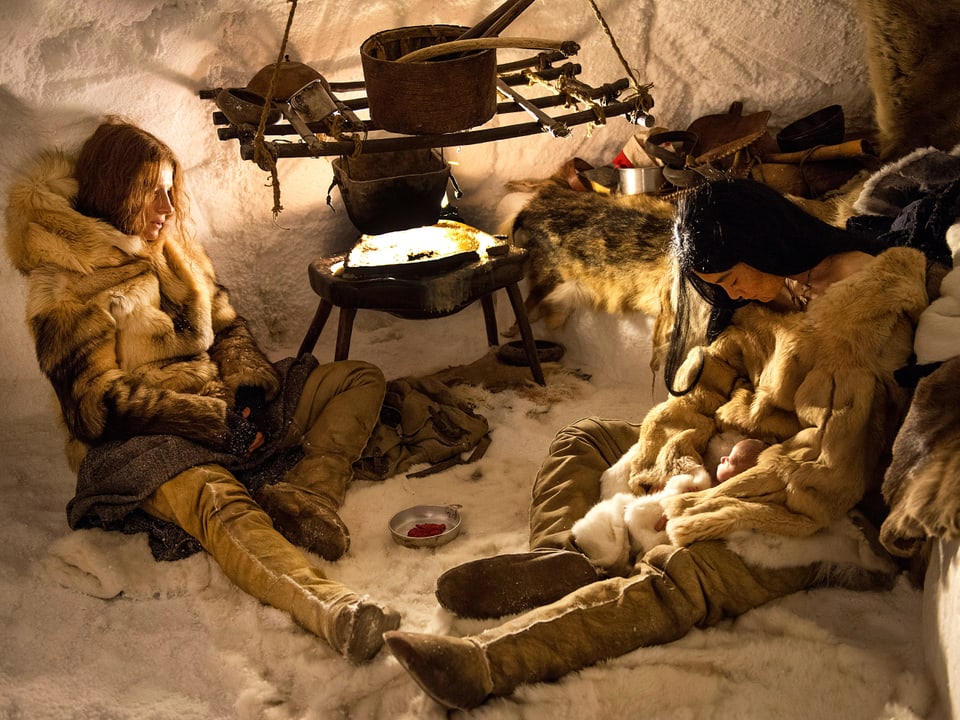 Die Frau des Polarforscherus und die Inuit sitzen in einem Iglu. Sie scheinen erschöpft. Die Inuit hält ein kleines Kind.