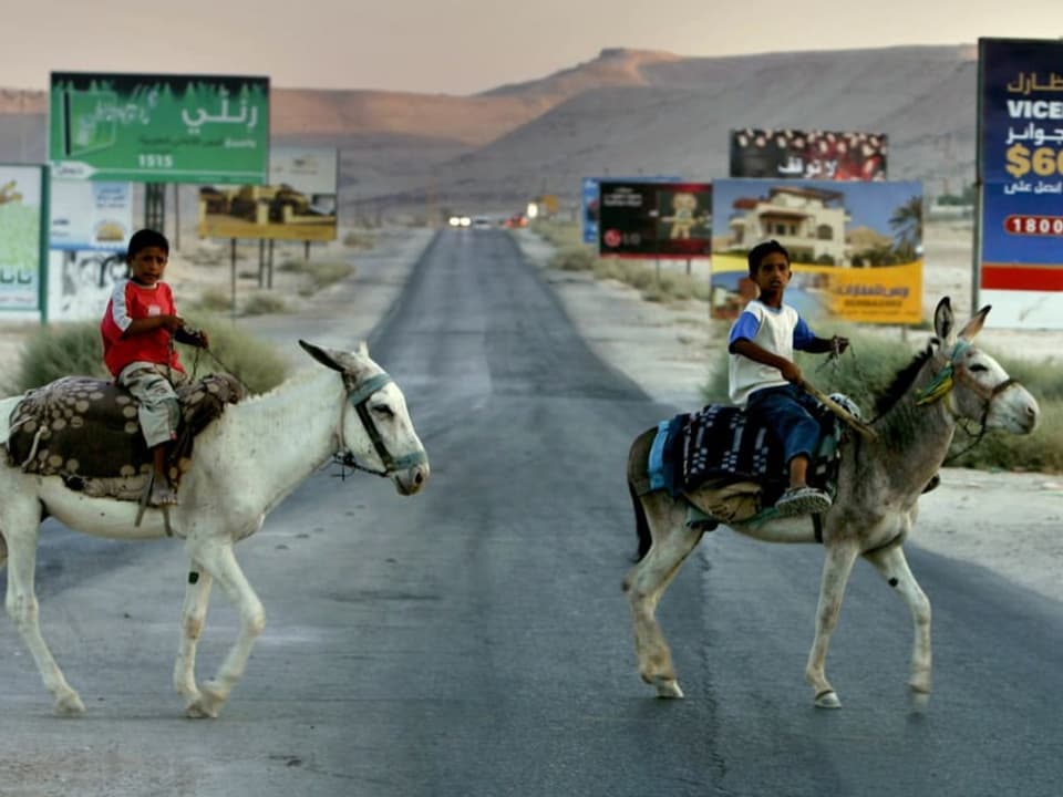 Zwei Kinder reiten auf ihren Eseln und überqueren eine Strasse.