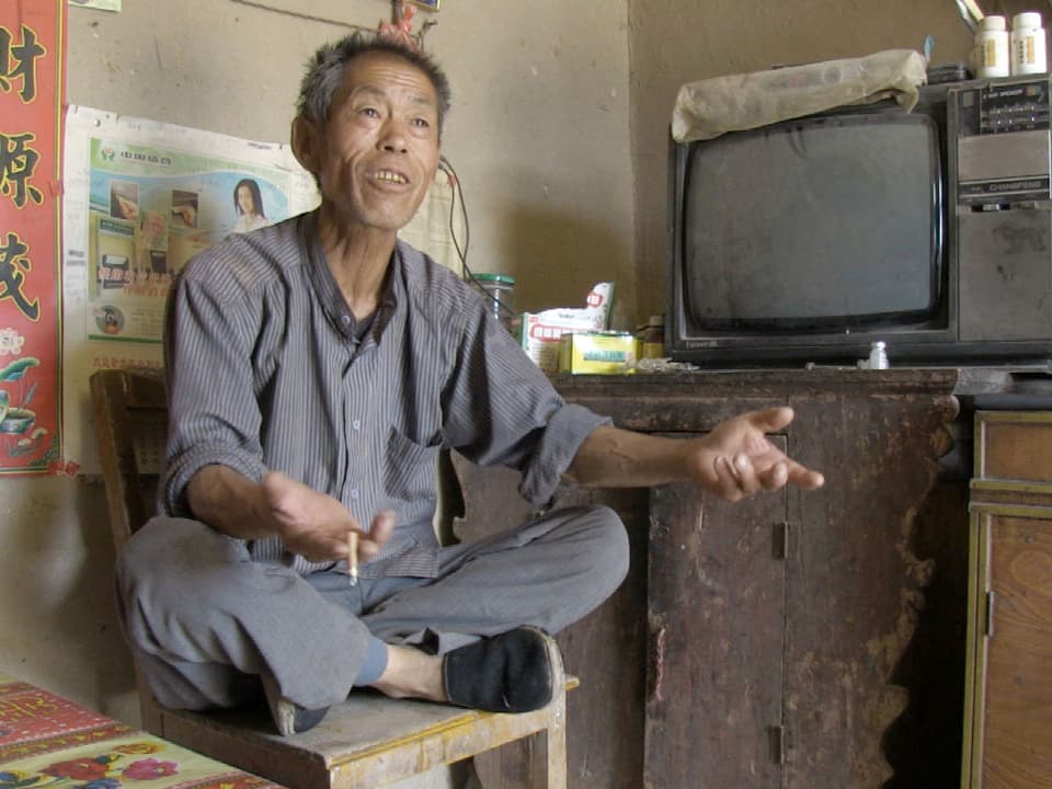 In einem staubigen und kargen Zimmer sitzt ein älterer chinesischer Mann im Schneidersitz auf einem Stuhl, sprechend, mit Zigarette in der Hand.