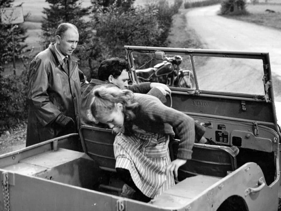 Ein Mann steht neben einem offenen Geländewagen und spricht mit dem Mann am Steuer. Ein Mädchen steigt gerade auf die Rückbank.