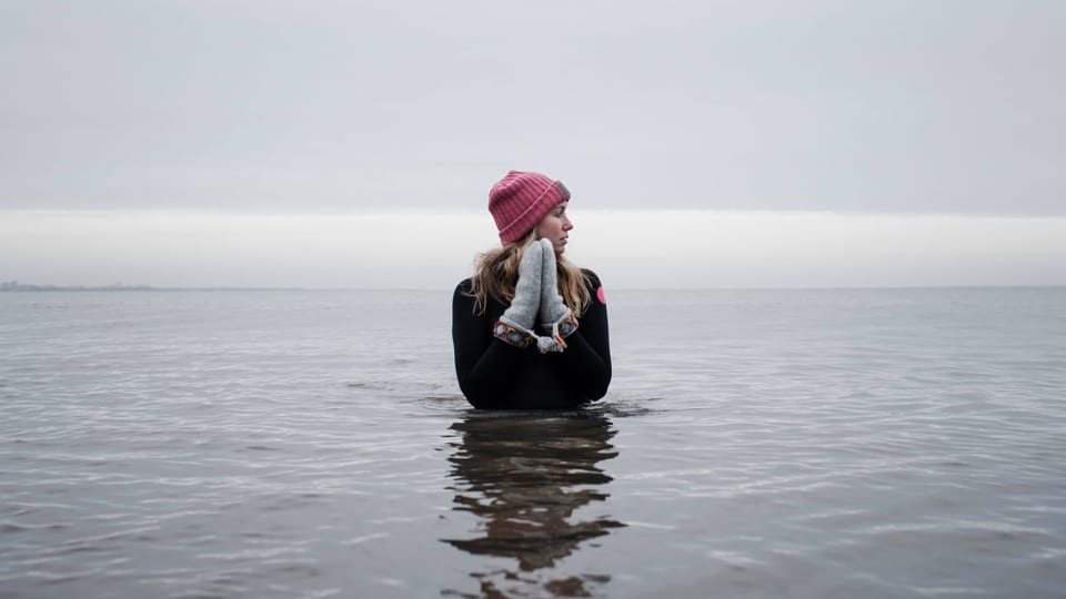 junge Frau steht mit Handschuhen und Mütze im Wasser, dahinter grauer Himmel
