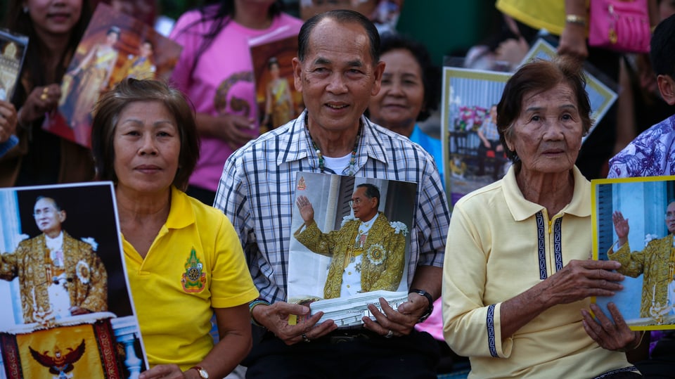 Thailänderinnen und Thailänder sitzen am Boden und halten Bilder von König Bhumibol in der Hand.