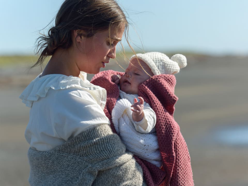 Eine Frau hält ein Baby in den Armen. Sie ist am Strand.