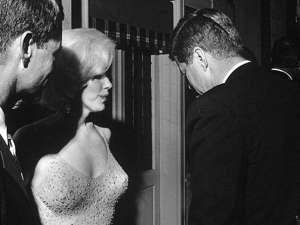 Schwarzweissbild: Mann in Anzug im Gespräch mit einer blonden Frau im Abendkleid.