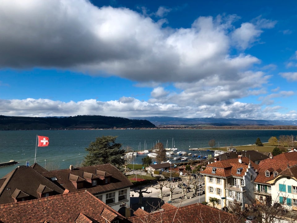 Von der Stadt Murten über die Dächer zum See. Eine Schweizflagge flattert auf einem Gäude. Der Himmel ist teils klar, teils von Wolken überzogen.