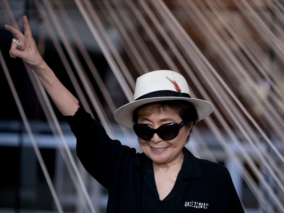 Yoko Ono strekt 2016 ihren Arm in die Luft. die Hand formt ein Peace Zeichen. 