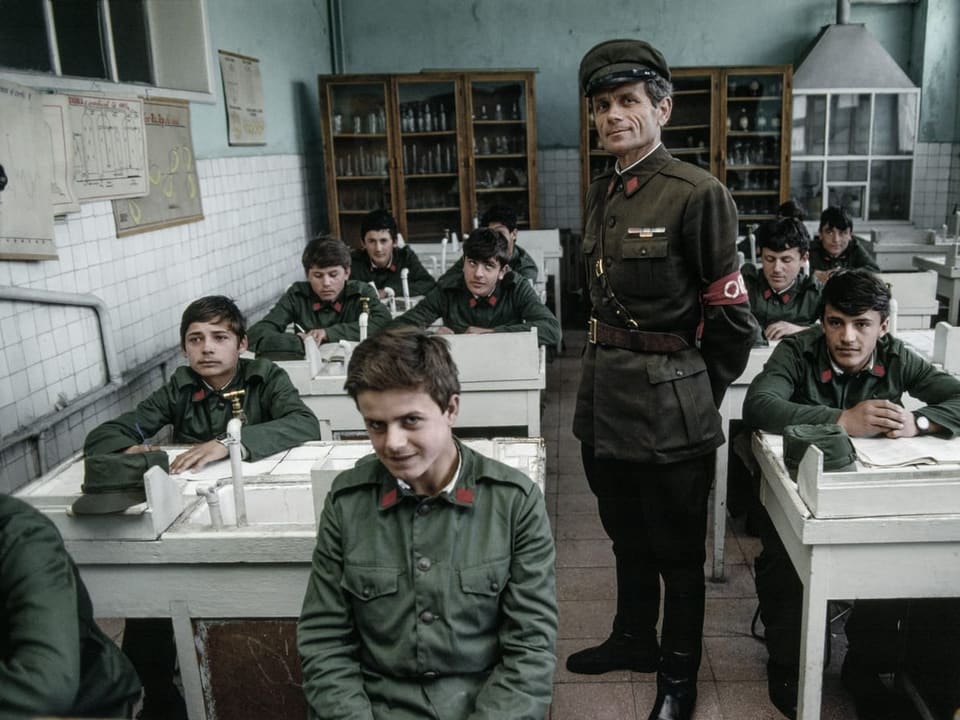 Kinder in Uniform sitzen in einem Schulzimmer, in der Mitte steht ein uniformierter Mann.