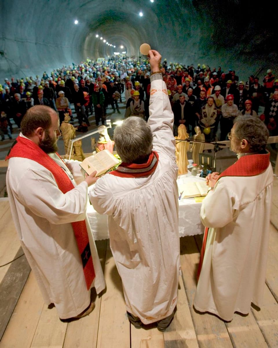 Drei Priester stehen im Tunnel, einer hält eine Hostie hoch.