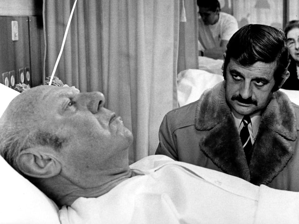 Ein alter Mann liegt im Spitalbett, daneben sitzt ein Mann mit Mantel und Schnurrbart.