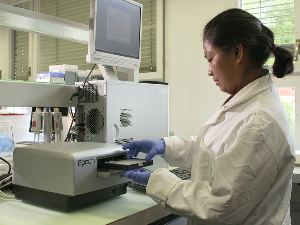 Frau in einem Labor mit weissem Kittel und blauen Schutzhandschuhen bedient ein technisches Messgerät.