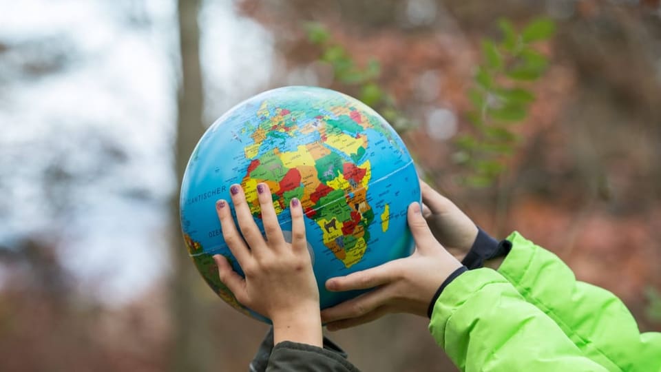 Vier Kinderhände halten in einem Wald einen farbigen Globus.