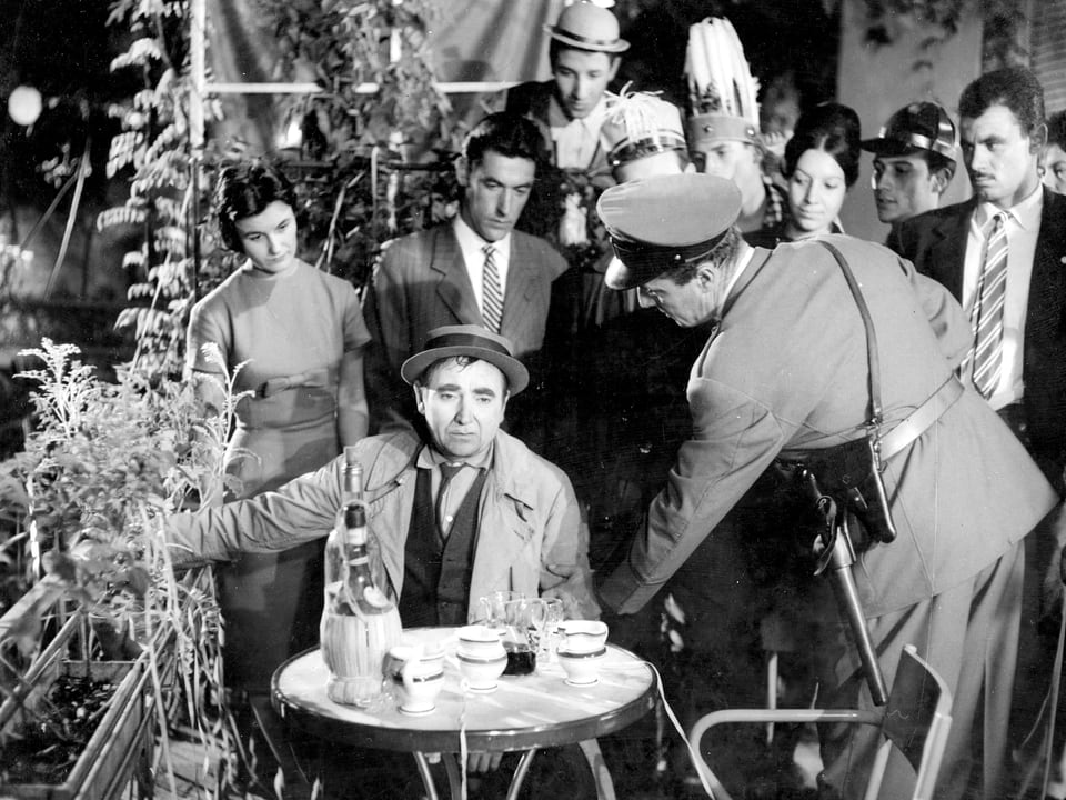 Ein Mann sitzt unglücklich an einem Kaffee-Tisch. Ein Polizist greift ihn am Arm. Viele Menschen stehen um ihn herum und beobachten die Szene.