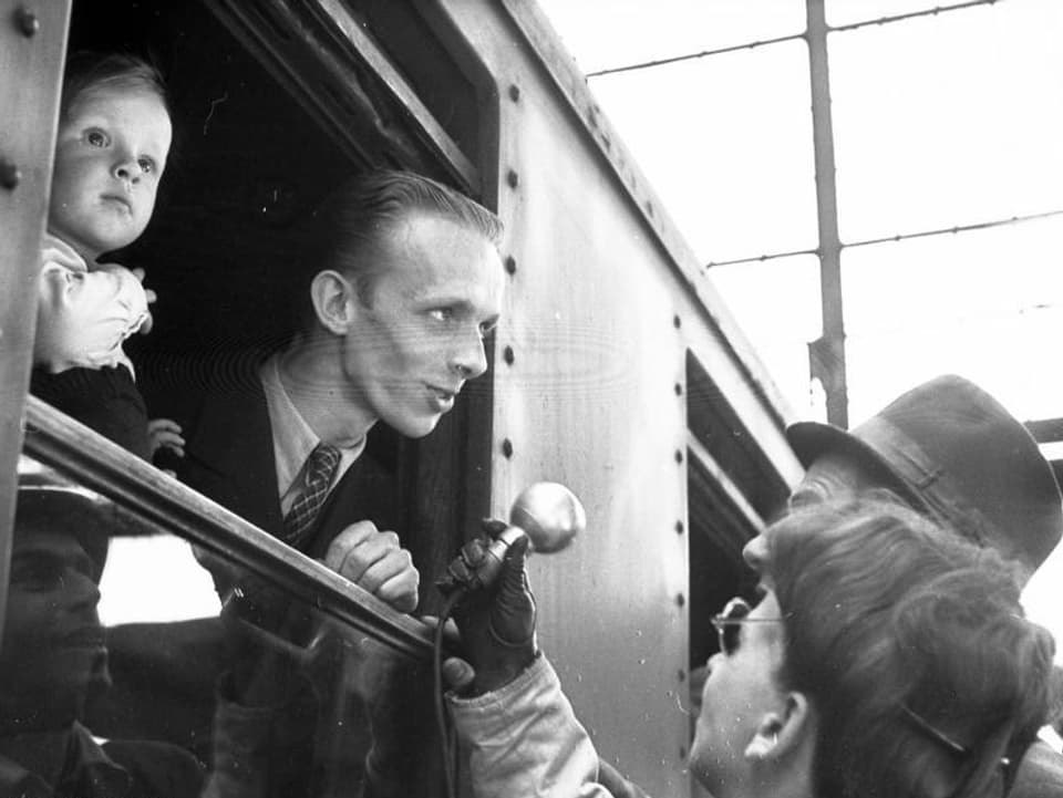 Ein Mann mit Kind auf dem Arm schaut aus dem Zug, ein Reporter streckt ihm ein Mikrofon hin.