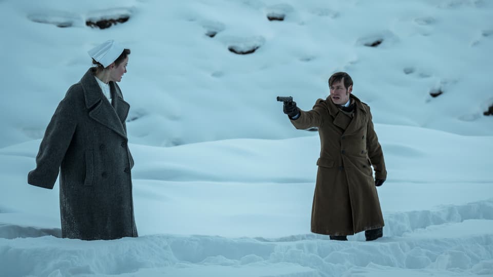 Frau im historischen Krankenschwester-Kostüm und ein Mann stehen im knietiefen Schnee. Der Mann bedroht sie mit Waffe. 