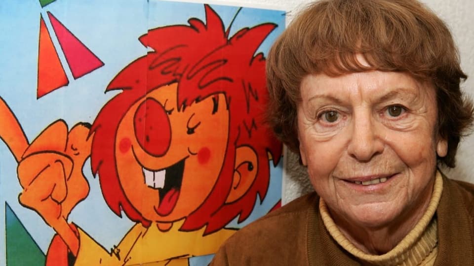 ältere Frau mit rötlichem Haar schaut in die Kamera, rechts ein Plakat mit rothaariger Comicfigur