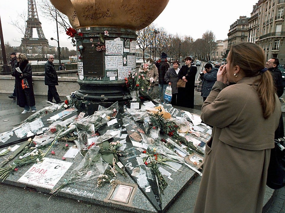 Zu sehen ist ein Platz in Paris in der Nähe des Eiffelturms. Passanten legen Blumen nieder.