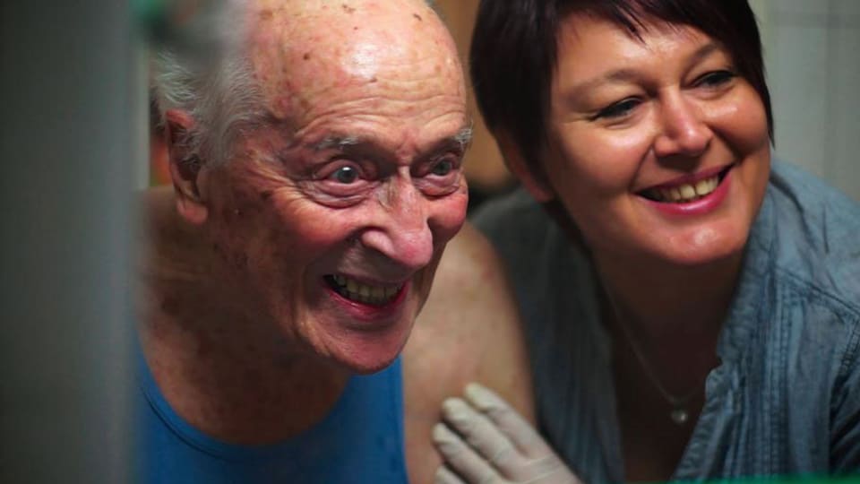 Ein älterer, pflegebedürftiger Mann, daneben seine Pflegerin, beide lachen.