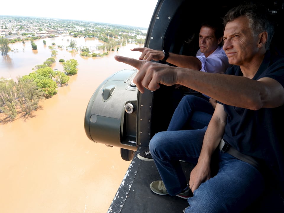 Argentinischer Präsident Mauricio Macri (vorne), hinter ihm ein anderer Mann, beide zeigen mit dem linken Arm aus dem offenen Heilkopter auf die Fluten.