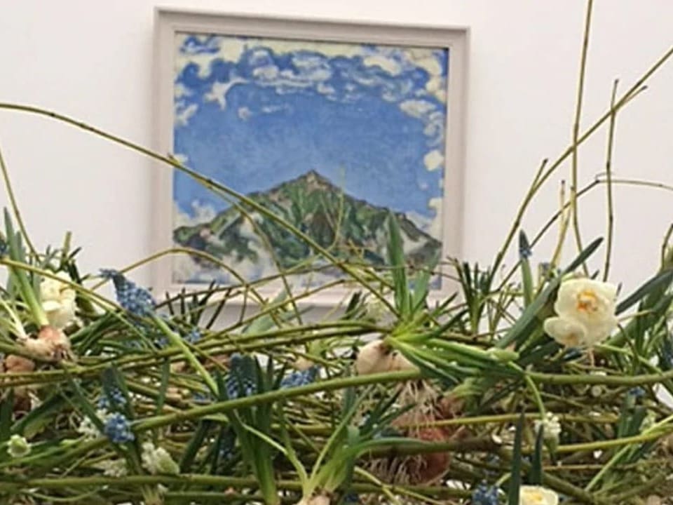 Ein Bild von einem Berg, davor ein Kranz aus Grünen Stängeln und weissen Blumen. 