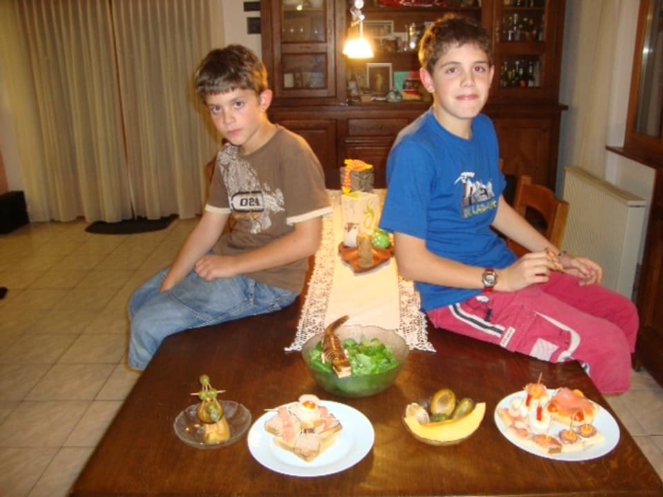 Zwei Söhne der Ambrosinis sitzen auf dem Esszimmertisch. Vor sich das zubereitete Essen.