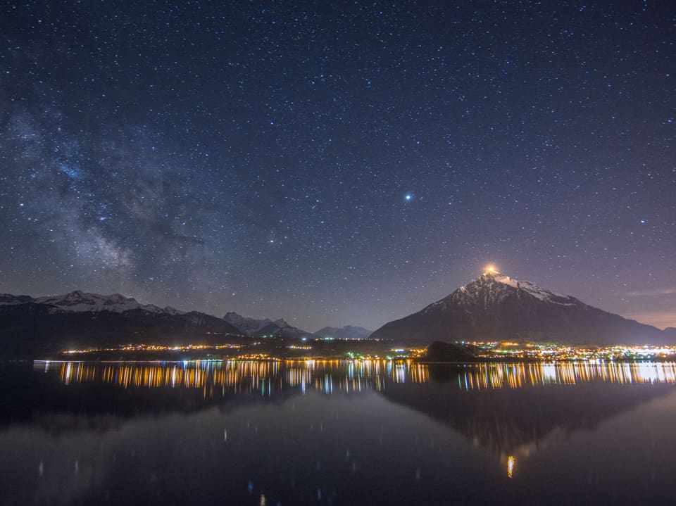 Nachtfoto mit Sternenhimmel und Lichtern von Häusern um einen See und einem hellen Licht auf einem Berg hinter dem See. 