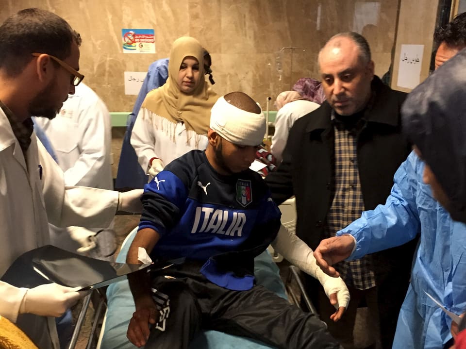 Spitalpersonal hilft einem Mann mit bandagiertem Kopf und eingebundenem linken Arm von einem Behandlungsbett aufstehen