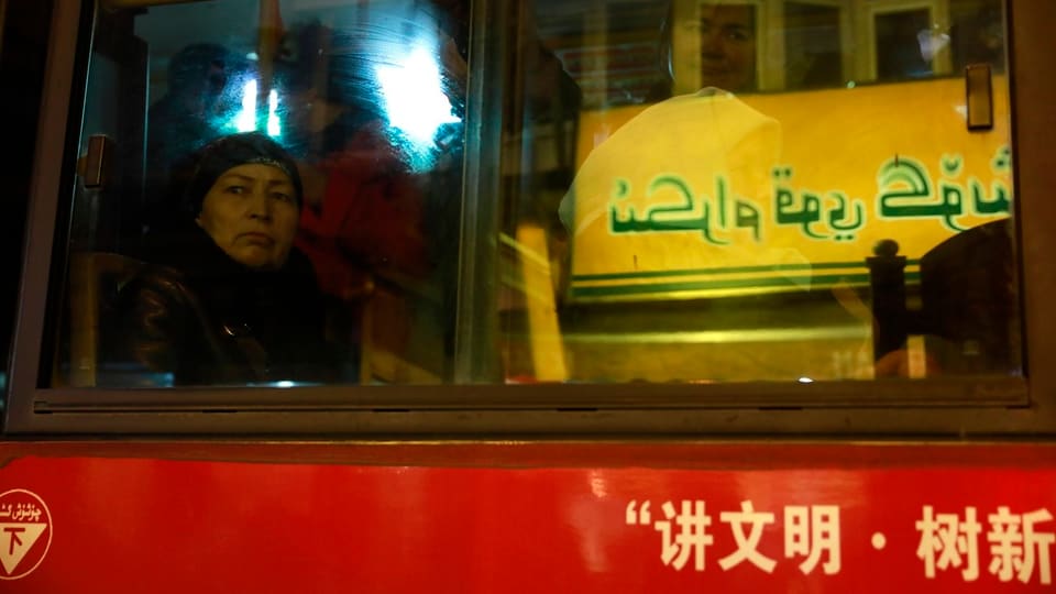 Eine Frau sitzt in einem Bus. Der Bus ist chinesisch beschriftet und fährt an einer Tafel mit arabischen Texten vorbei.