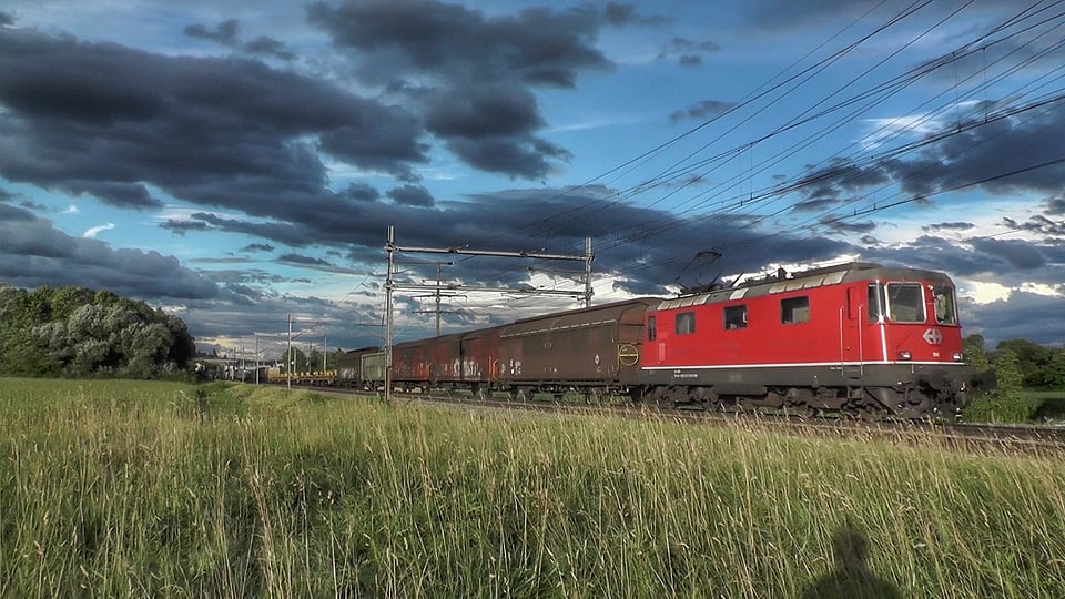 Ein Zug mit einer roten Lok fährt über eine Wiese. Am Himmel stehen Wolken in allen Grautönen.