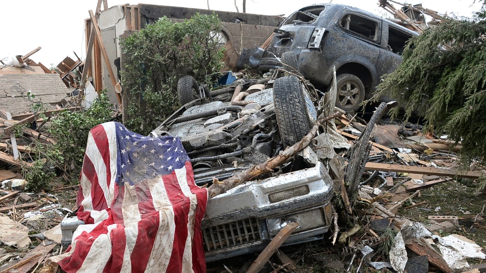 Häuser liegen auf Trümmerhaufen. Im Vordergrund eine US-Flagge.