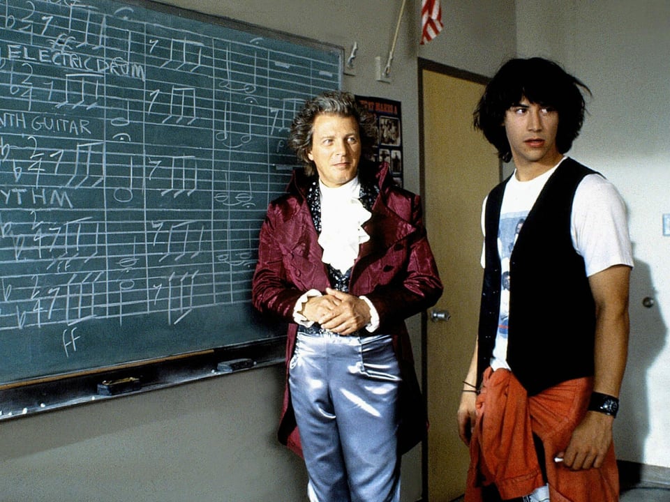 Zwei Männer vor einer mit Noten beschriebenen Wandtafel. Der eine Mann soll Beethoven nachstellen mit alter Kleiderung. Rechts von ihm ein junger Mann.