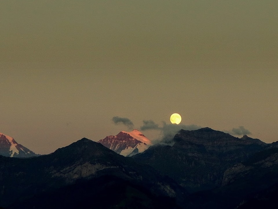 Konturen von Bergen im Vordergrund, über den Schneebergen ein kleiner, voller Mond. 