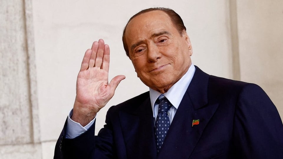 Silvio Berlusconi winkt in die Kamera. Anzug, blaue Krawatte.
