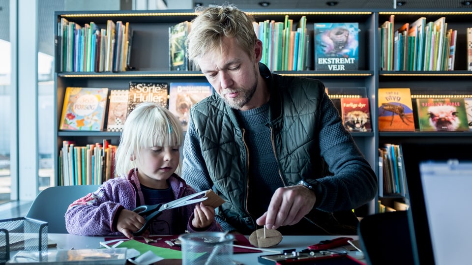 Ein MAnn und ein Kind sitzen nebeneinander an einem Tisch in einer Bibliothek und bastel mit Schere und Karton.