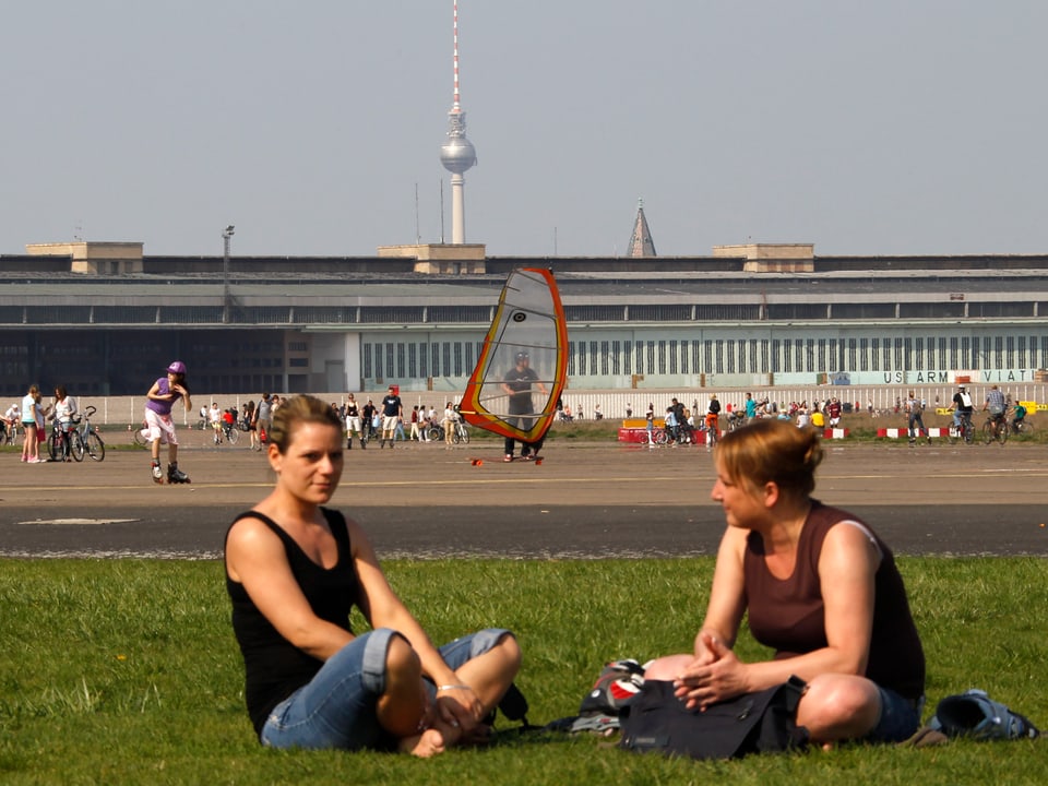 Im Vordergrund sitzen zwei Frauen auf dem Rasen und sprechen miteinander, im Hintergrund Skater und Windsurfer auf der alten Landebahn.