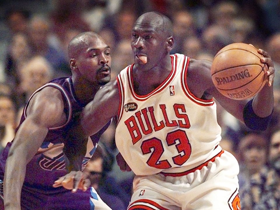 Michael Jordan spielt mit einem Basketball in der linken Hand im Bulls-Trikot gegen einen gegnerischen Spieler.