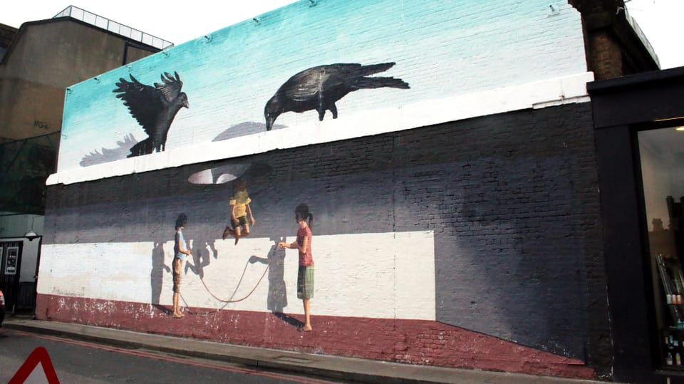 Wandmalerei: Seilspringende Kinder. Darüber ein Dach mit einem Loch. Darauf sitzen zwei Krähen und schauen durch das Loch. 