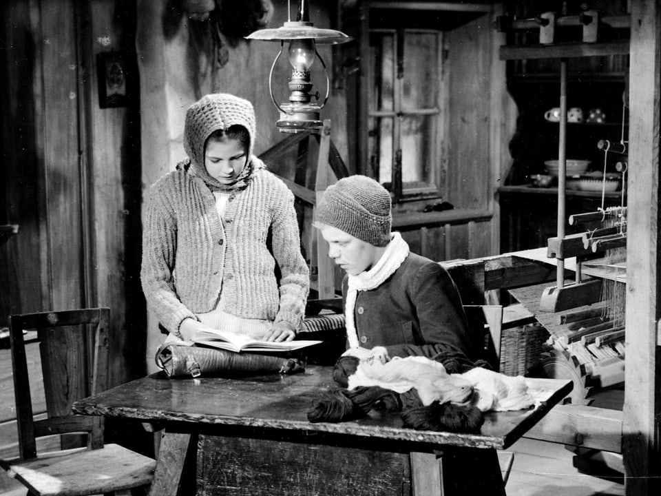 Heidi und Peter an einem Tisch in Winterkleidung. Heidi stehend, liest von einem Blatt Papier.