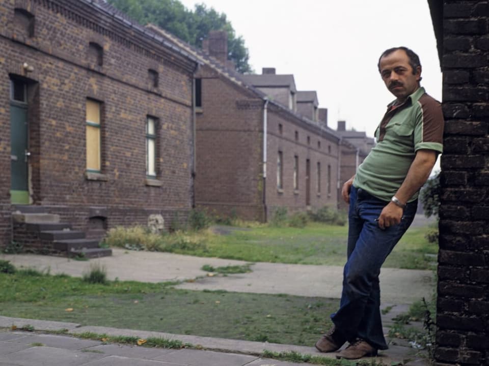 Ein Mann lehnt an eine Ziegelwand, in Hintergrund weitere Ziegelgebäude