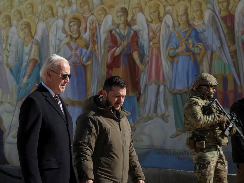 Zwei Männer gehen vor einer bunt, mit heiligen Fresken bemalte Wand. Rechts steht ein bewaffneter Soldat.