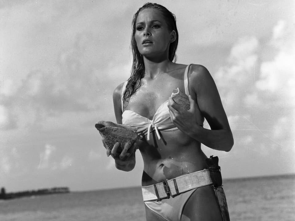 Eine blonde Frau im Bikini hält eine Muschel in der Hand und steht knöcheltief im Meer.