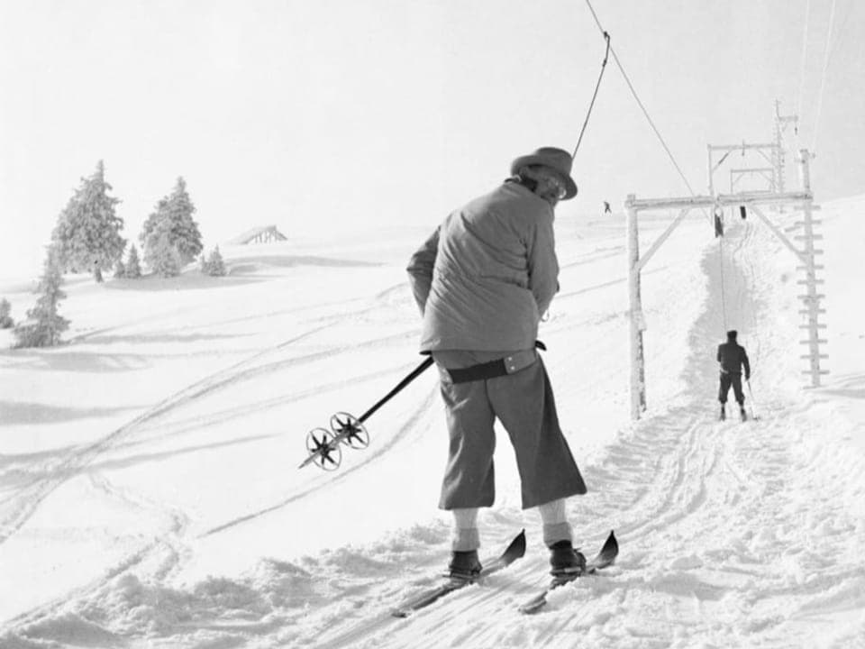 schwarzweiss Foto, Mann mit Stöcken auf Skiern an Bügellift, Rücken zum Fotograf, schaut zurück.