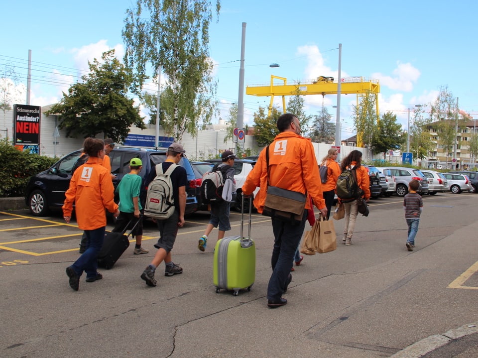 Familie Ambrosini und Familie Leonardi marschieren mit ihrem Gepäck und in orangen Jacken über einen Parkplatz.