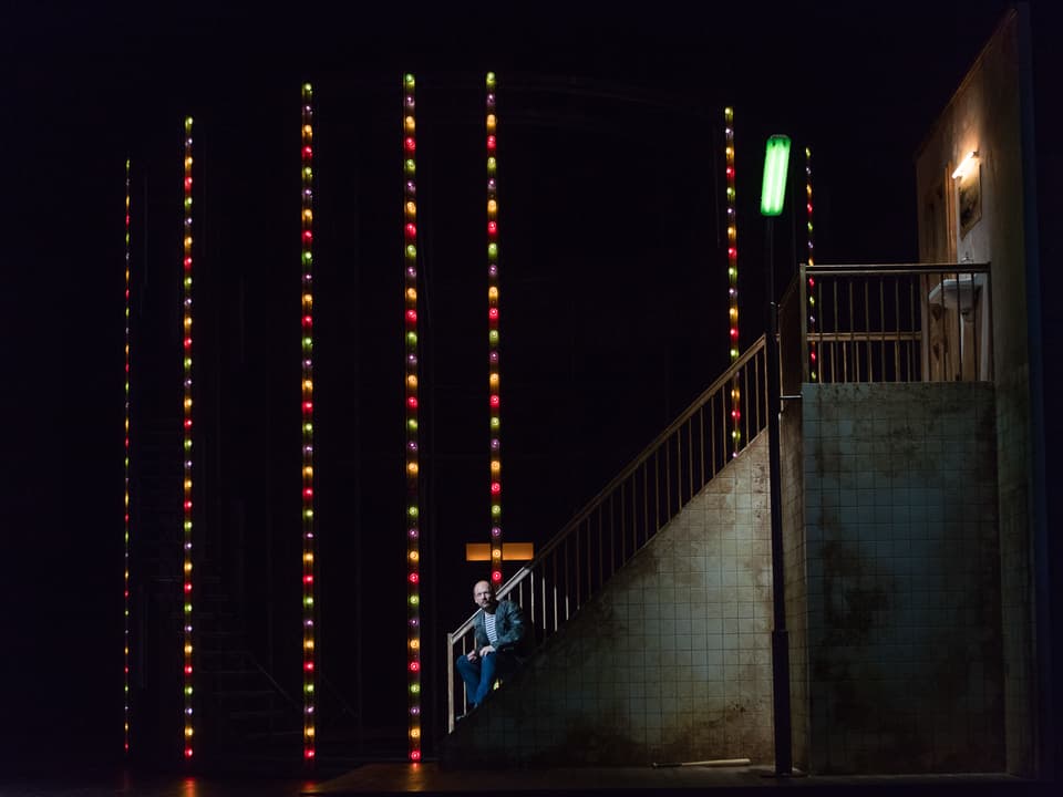 Ein Mann sitzt irgendwo in der Dunkelheit, von oben herab hängen Lichterketten mit verschiedenen farbigen Lämpchen.