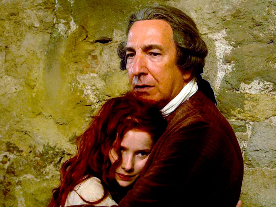 Ein Mann mit langhaariger Perücke hält ein Mädchen in seinen Armen.